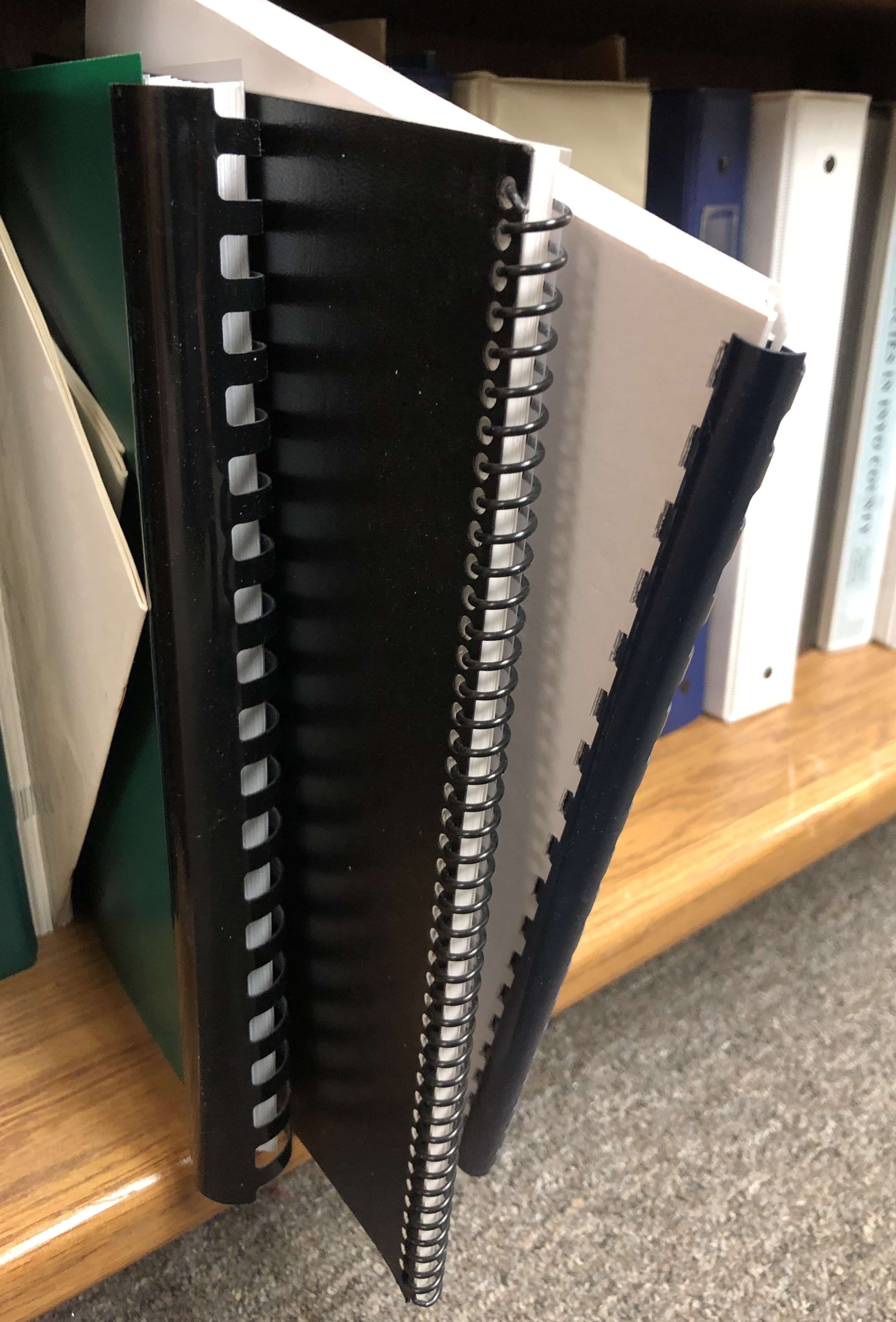 Multi-sized spiral-bound reports sticking out from bottom level of book shelf.
