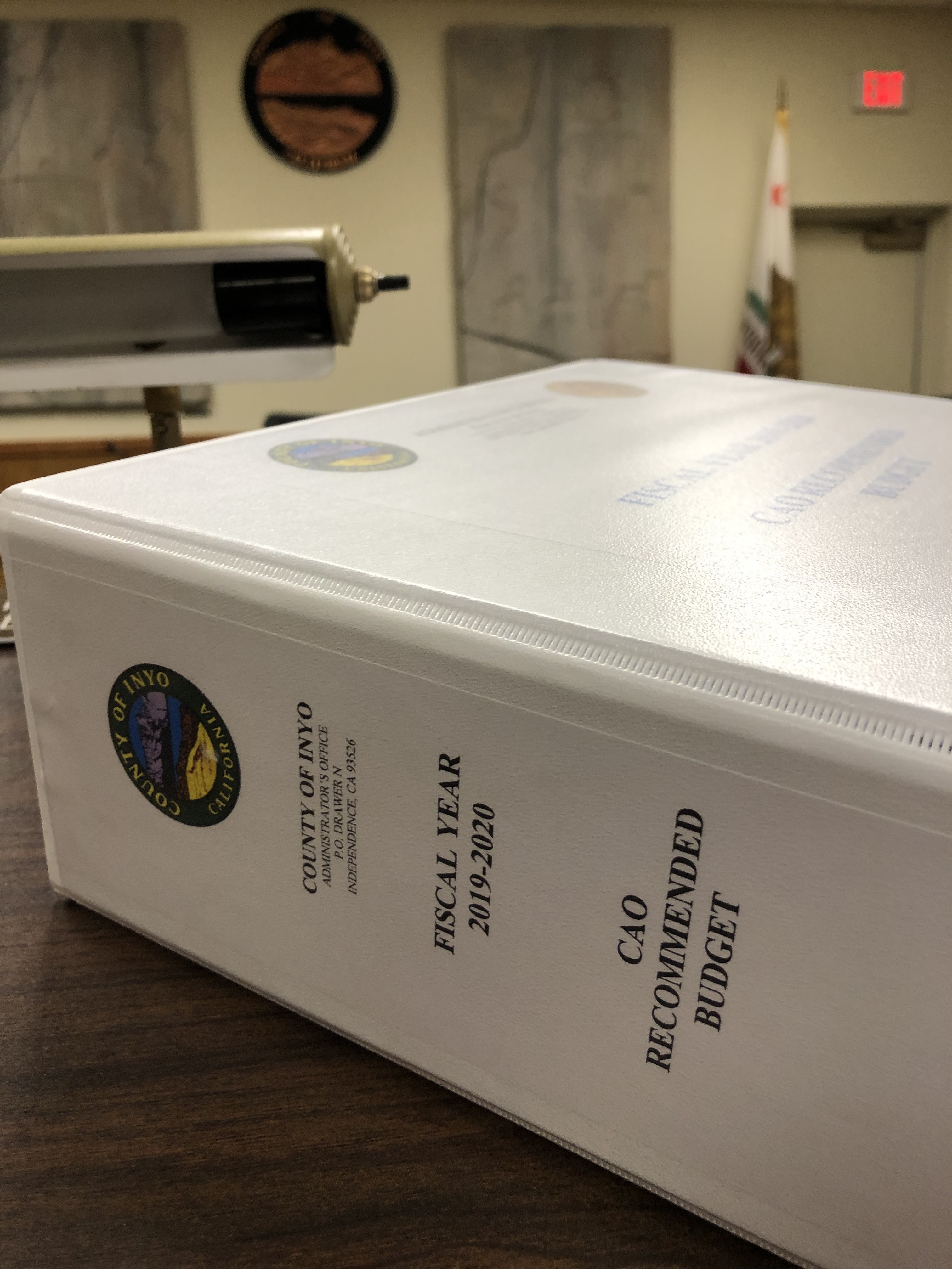 Three-inch thick white binder sitting atop podium in Board Room with County Seal on wall in background.