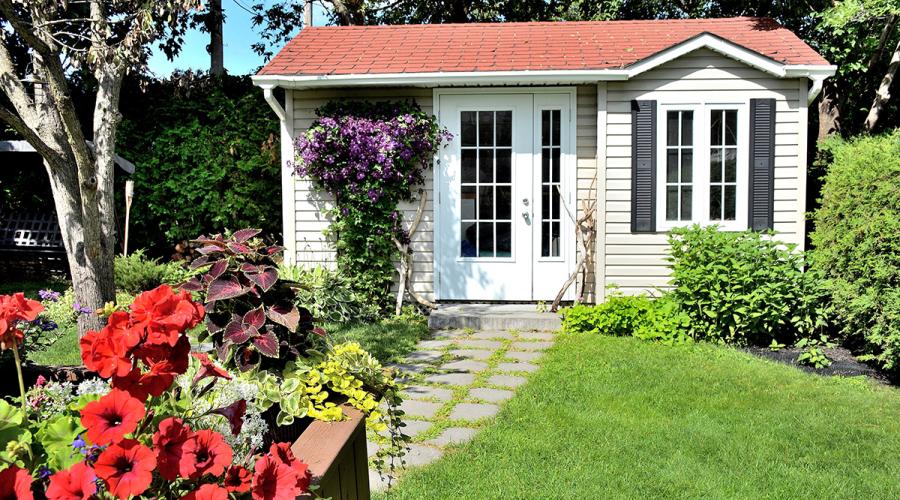 Small, white wooden home in backyard with green grass and lots of flowers.