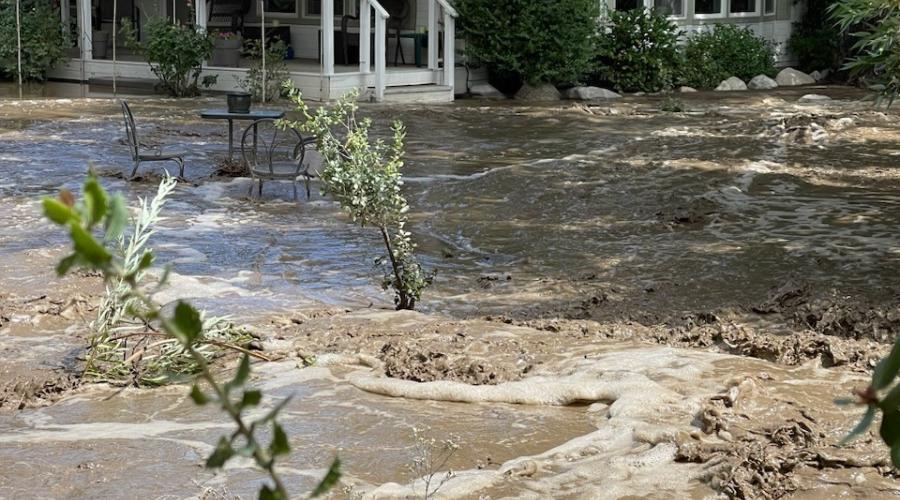 White mobile home surrounded by muddy, fast-moving flood water.