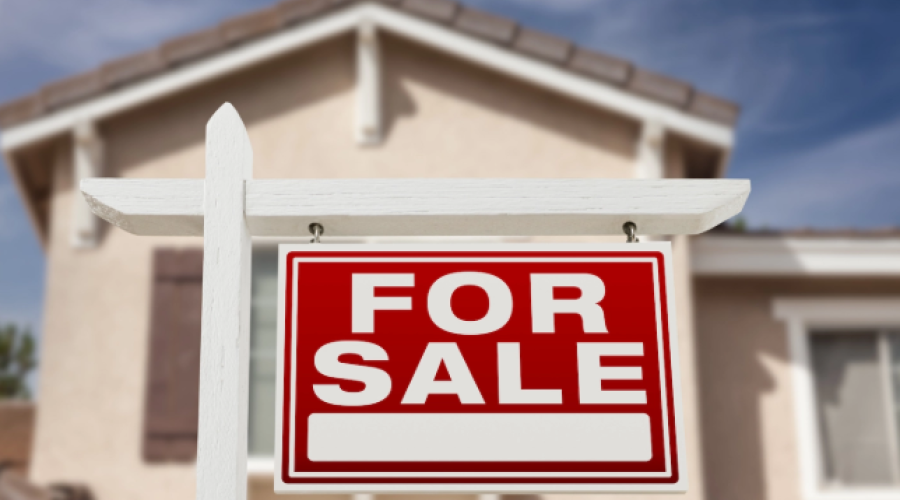 A red For Sale sign with white lettering hanging from a wooden post with a cream-colored house in the background.