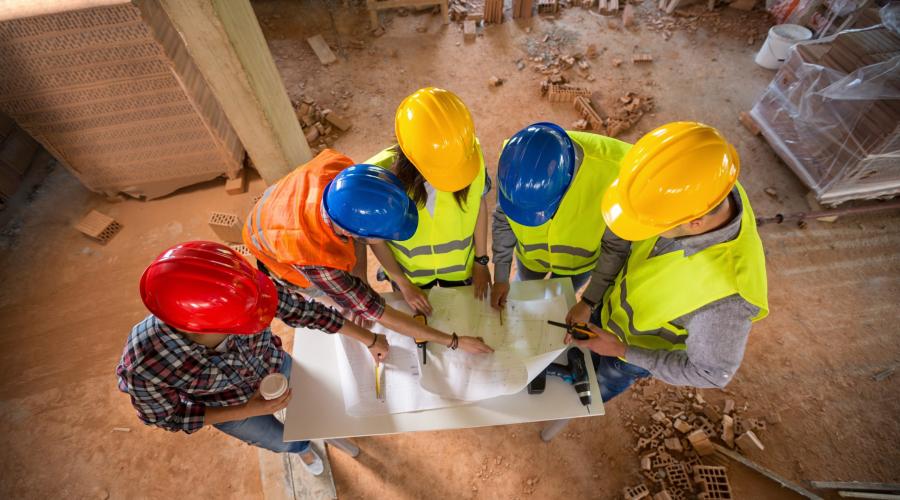 Five individuals in different-colored hard hats are seen from above poring over blueprints at a construction site.