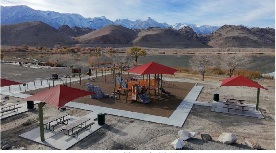 Drone view of a playground and shaded picnic area with a lake, rolling hills, and jagged peaks in the background.