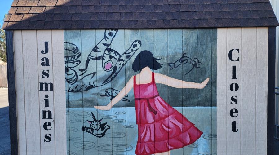 Mural on side of shed with the words Jasmine's on the left side of the panel and the word Closet on the right side, depicting a female, shown from the back, with dark hair and a magenta dress dancing in front of images of a cat, a tiger, and perhaps a sheep or lamb.