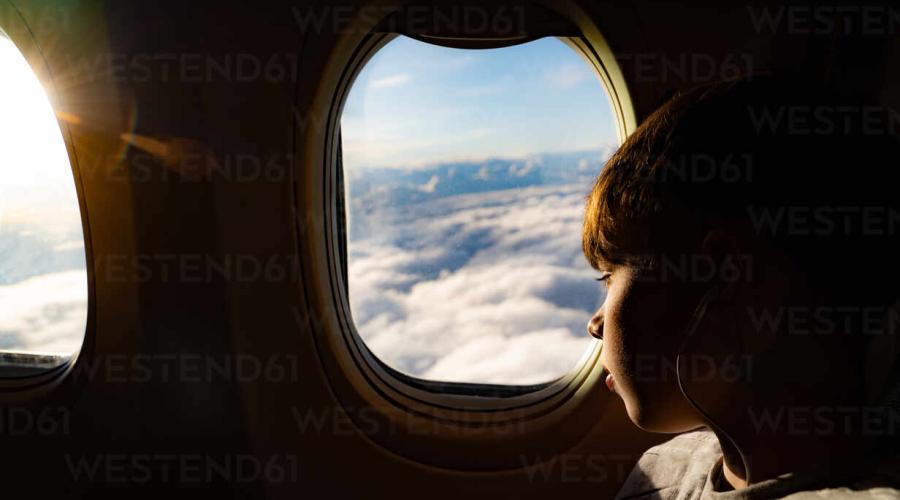 Young boy, in profile, looking out an airplane window at clouds and blue sky.