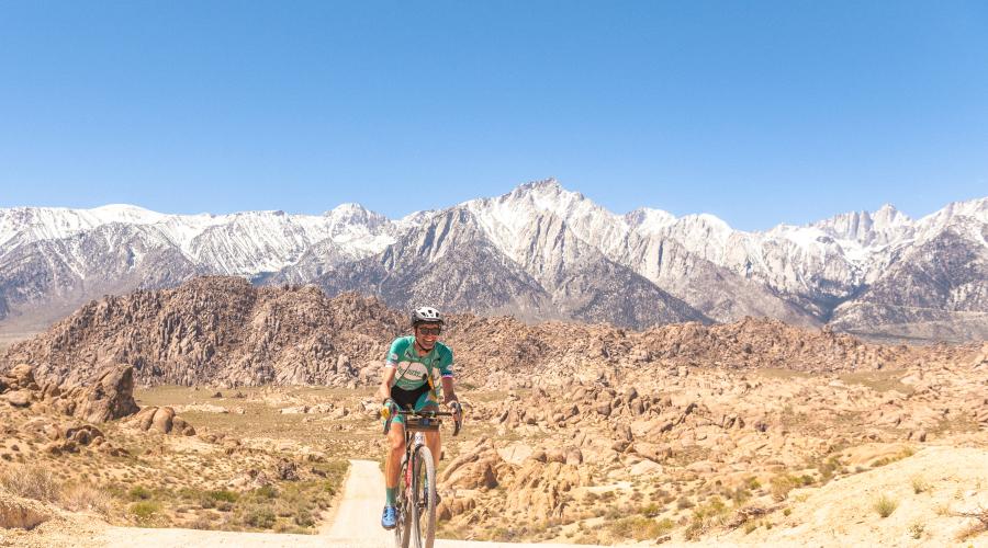 A cyclist in green lycra is pedaling uphill on a dirt road with the Sierra Nevada in the background