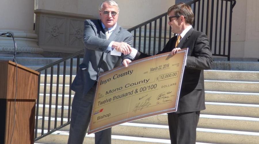 Two men in suits on courthouse steps shake hands over giant novelty check