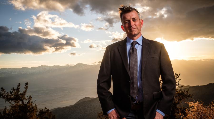 Man in suit standing in front of sunset in forest.