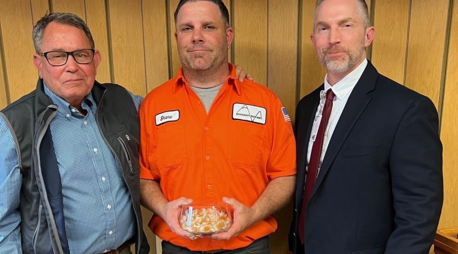 Three men standing against wood-paneled wall, one in middle holding a candy dish
