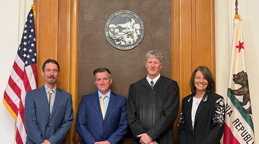 Two men in suits, a robed judge, and a woman standing between the U.S. and California state flags.